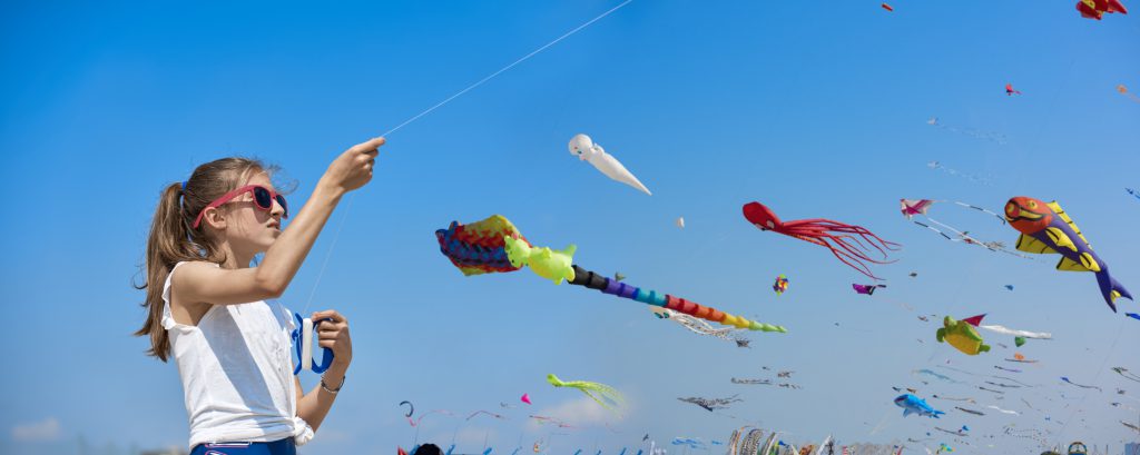 The little girl in the t-shirt plays on the beach by the sea with a colorful kite. In the background many people are walking on the beach.