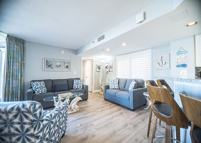 Picture of the inside of one of our condo's we offer at Sea Watch Resort. Couch, loveseat, chairs, barstools, pictures, hardwood floors
