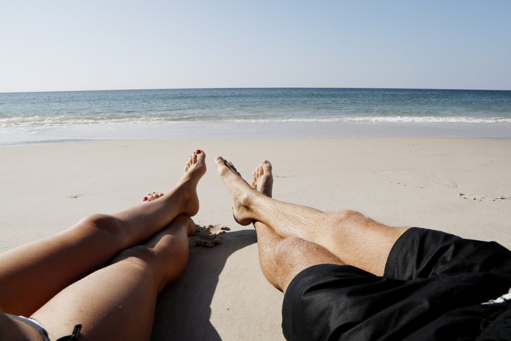 a couple, man and woman sit on an empty beach looking out onto the ocean from the sand with their legs crossed
