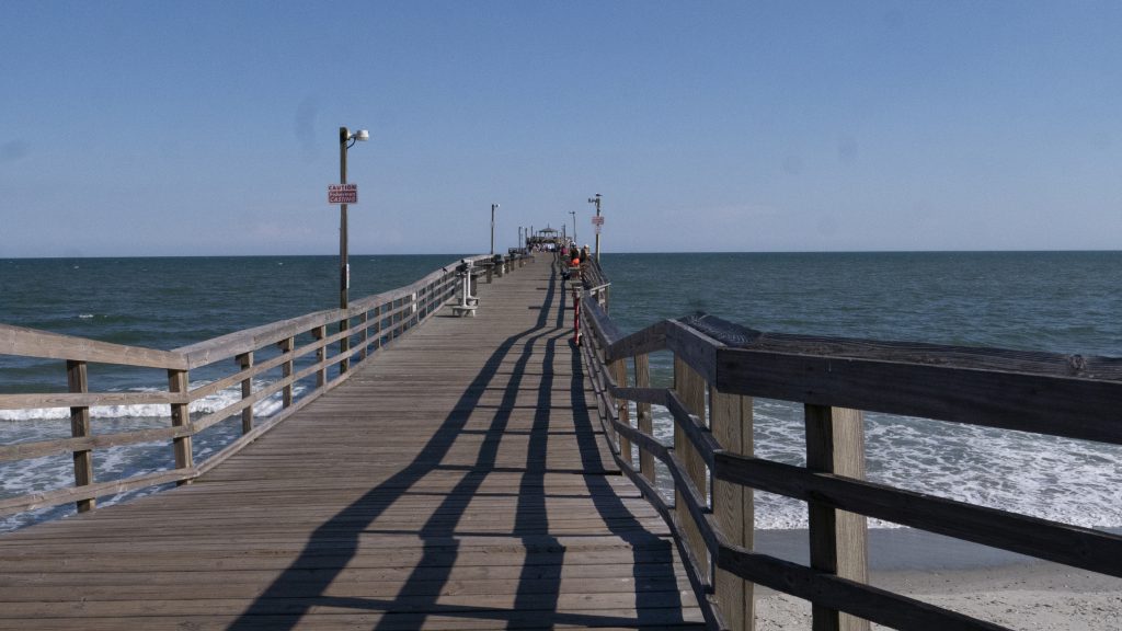 wooden pier with people fishing and walking on it with the blue sky, and ocean and sand