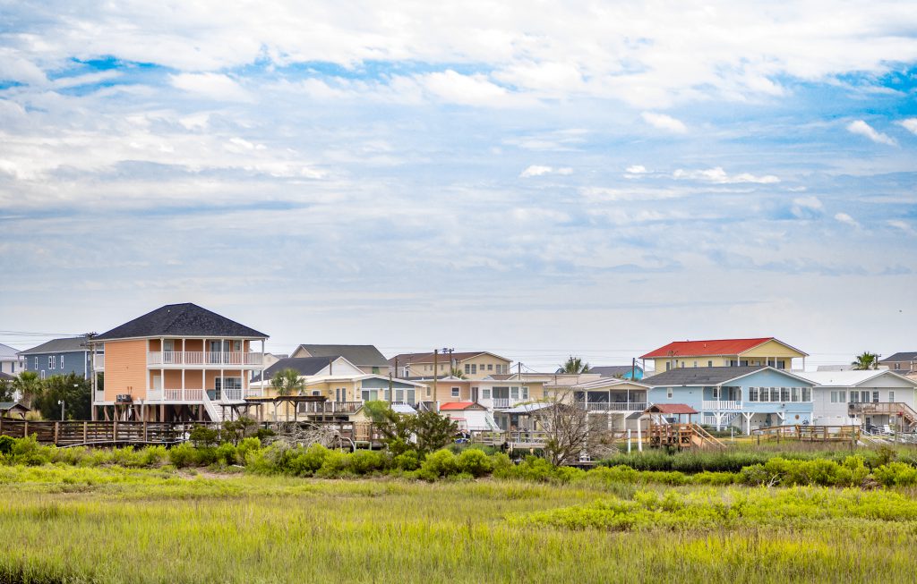 A row of colorful coastal vacation houses near the sea built for tourists in the North Myrtle Beach, South Carolina. Grass in a marsh in the foreground and dramatic overcast sky in the background.