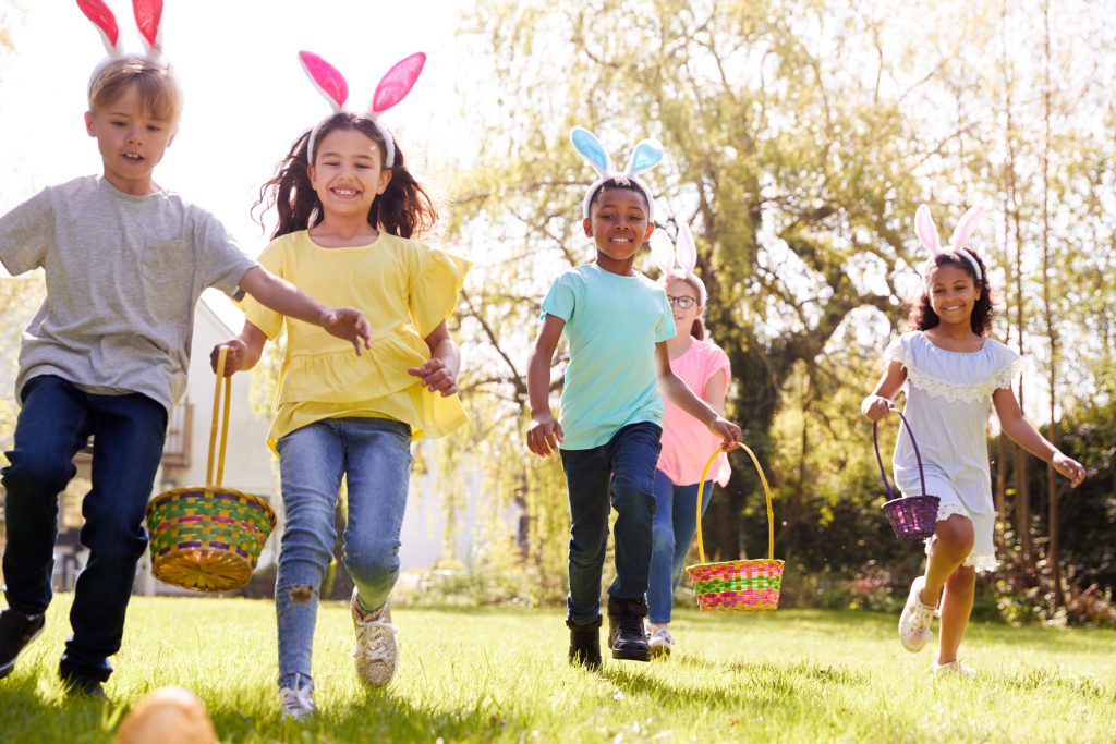 Group Of Children Wearing Bunny Ears Running To Pick Up Chocolate Egg On Easter Egg Hunt In Garden