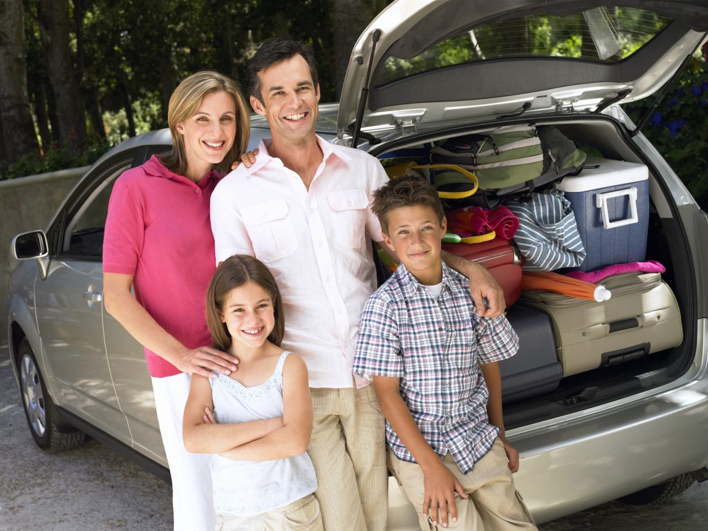a family with a mom, dad, son and daughter standing in front of their packed car for their beach vacation
